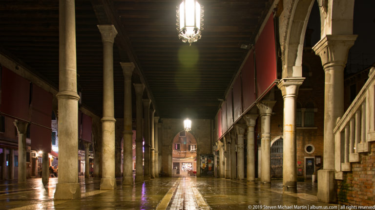 Deep colonnaded arcade with wet paving under nighttime illumination in Venice.