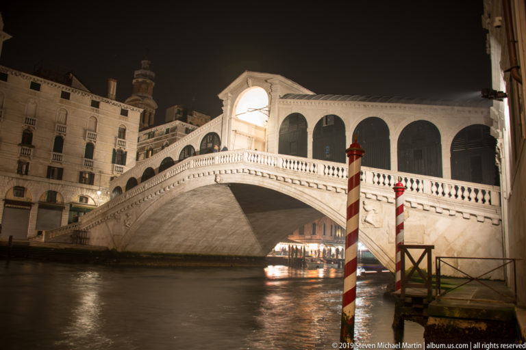 Rialto Bridge with striped mooring pole under nighttime illumination in Venice.