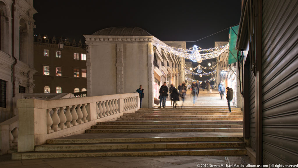 Pedestrians ascending the Rialto Bridge at night under nighttime illumination in Venice.
