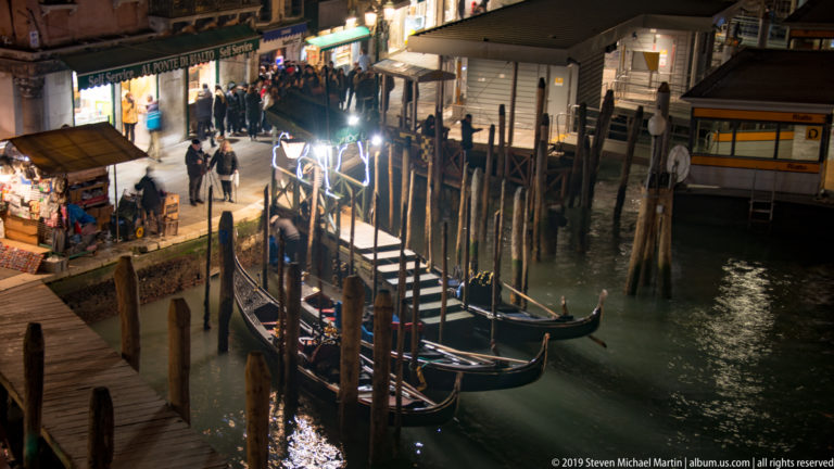 Gondolas moored beside a busy waterfront landing under nighttime illumination in Venice.