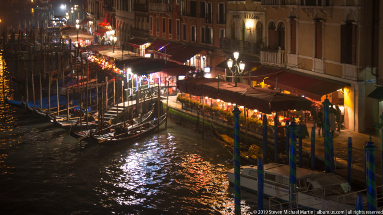 Gondolas beside illuminated waterfront awnings under nighttime illumination in Venice.