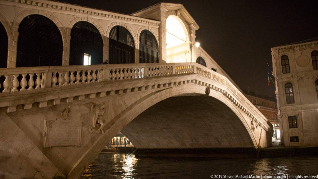 Illuminated Rialto Bridge rising over the dark Grand Canal, seen from a low waterside angle.
