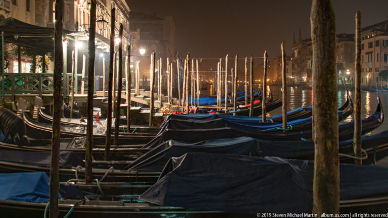 Covered gondolas packed among wooden mooring posts under nighttime illumination in Venice.