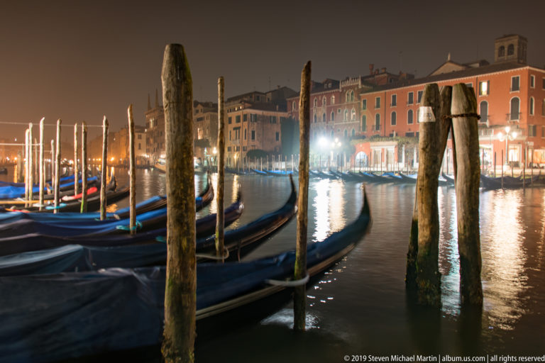 Gondolas and weathered posts with canal-front buildings beyond under nighttime illumination in Venice.