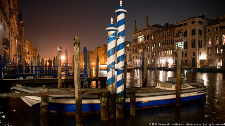 Blue-and-white striped mooring posts and small boat under nighttime illumination in Venice.
