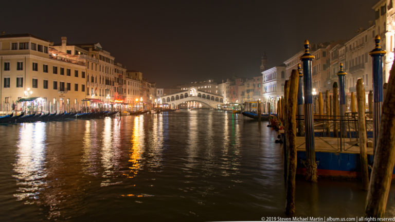 Wide nighttime view toward the Rialto Bridge under nighttime illumination in Venice.