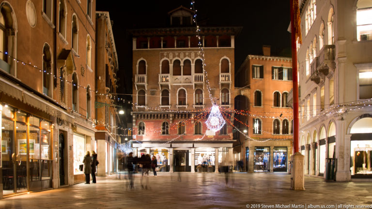 Small Venetian campo with festive lighting and shops under nighttime illumination in Venice.