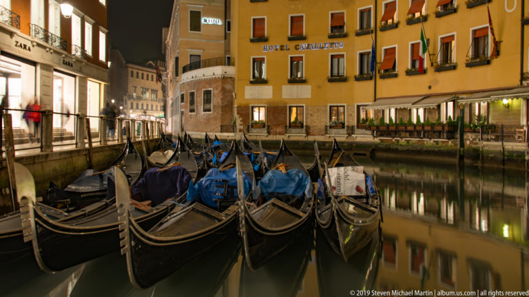 Black gondolas with blue covers moored on a calm Venetian canal beside the illuminated Hotel Cavalletto at night.