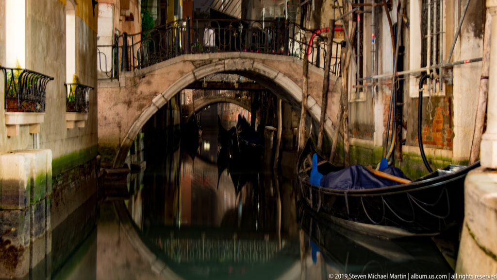Black gondola beside a narrow canal passing beneath two arched pedestrian bridges between closely spaced Venetian buildings.