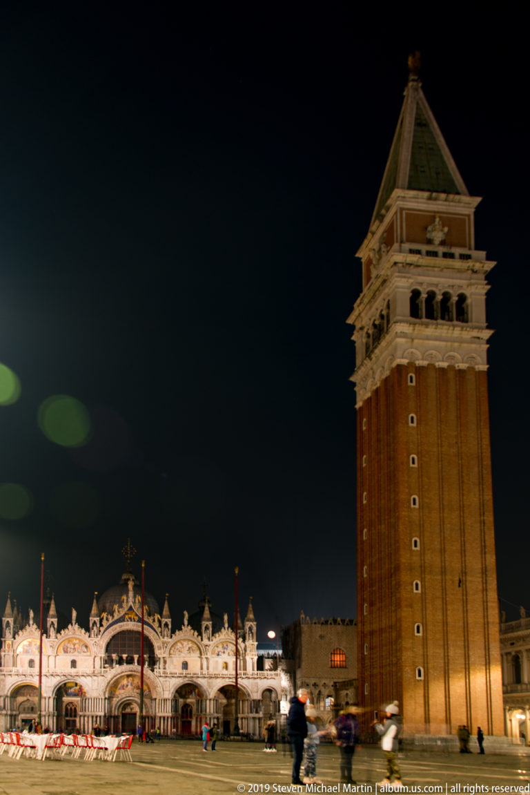 St. Mark's Campanile rising above illuminated arcades and small figures in Piazza San Marco at night.