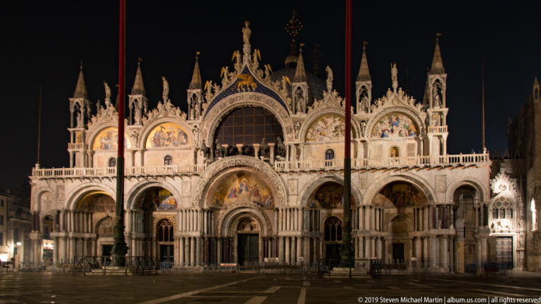 Illuminated façade of St. Mark's Basilica with arched portals, mosaics, columns, and ornate roofline against the night sky.