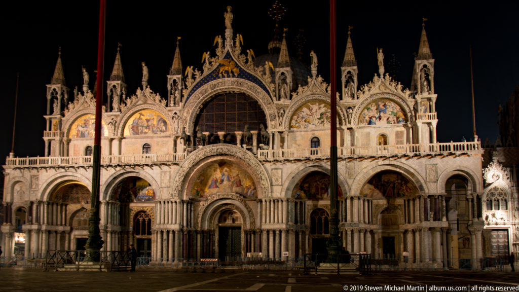 Closer nighttime view of St. Mark's Basilica, showing illuminated arches, mosaic lunettes, clustered columns, and roofline sculpture.