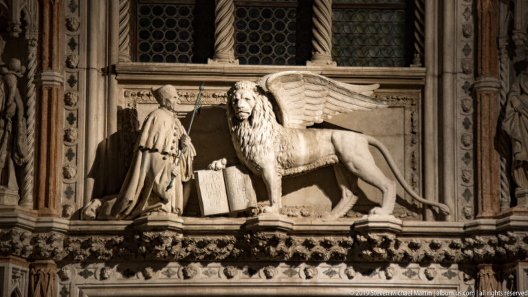 Winged Lion of St. Mark carved in stone beside a kneeling robed figure and open book, lit at night.