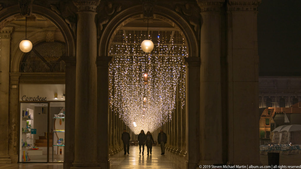 Pedestrians walking through a long arched arcade beneath dense cascading strings of white decorative lights.