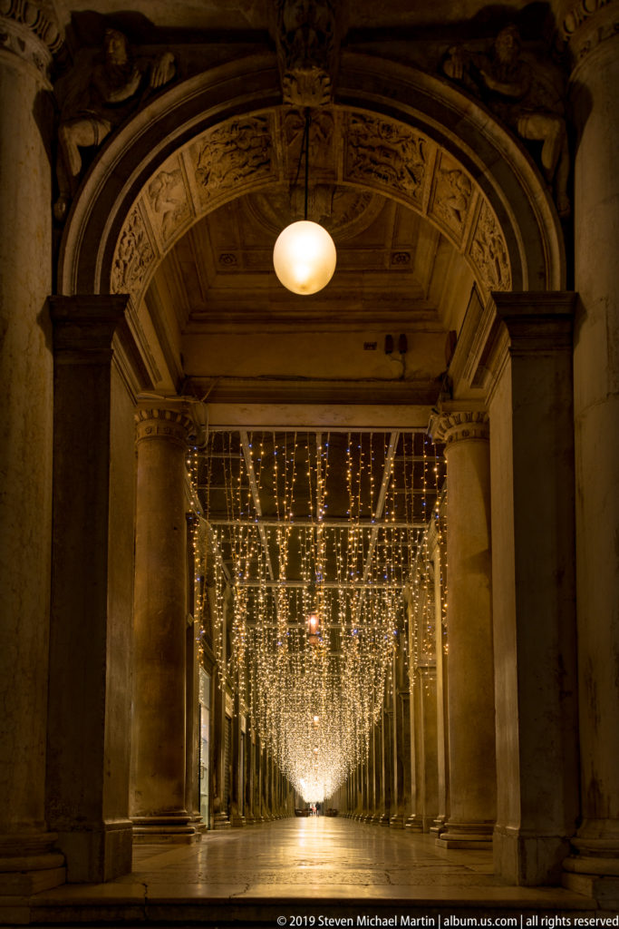 Centered view through a stone arcade with carved arch, columns, reflective pavement, and a dense canopy of white lights.