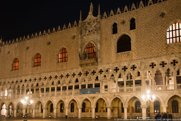 Illuminated Gothic façade of the Doge’s Palace with arcades and patterned upper walls against a black night sky.