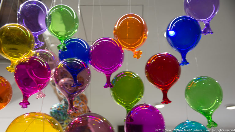 Cluster of brightly colored blown-glass balloon ornaments hanging under white shop lights.