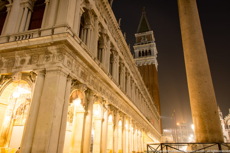 Illuminated Procuratie arcade receding toward St. Mark's Campanile, with a large stone column in the foreground.