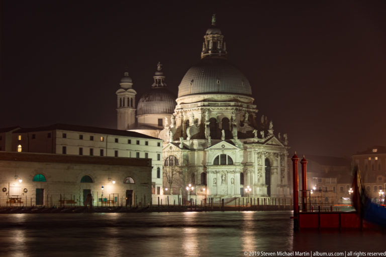 Illuminated Santa Maria della Salute with large dome and façade reflected across the Grand Canal at night.