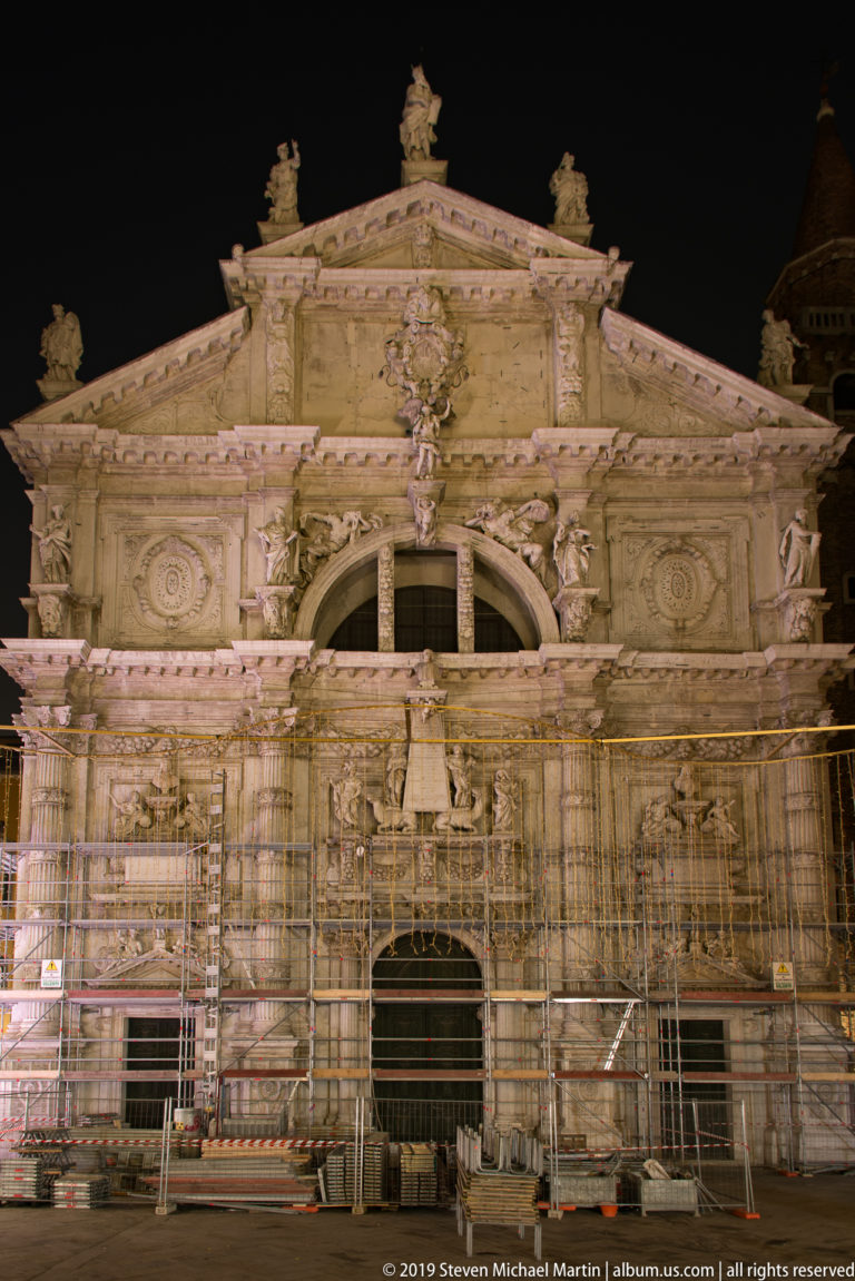 Ornate stone church façade with columns, statues, pediment, and extensive metal scaffolding during nighttime restoration.