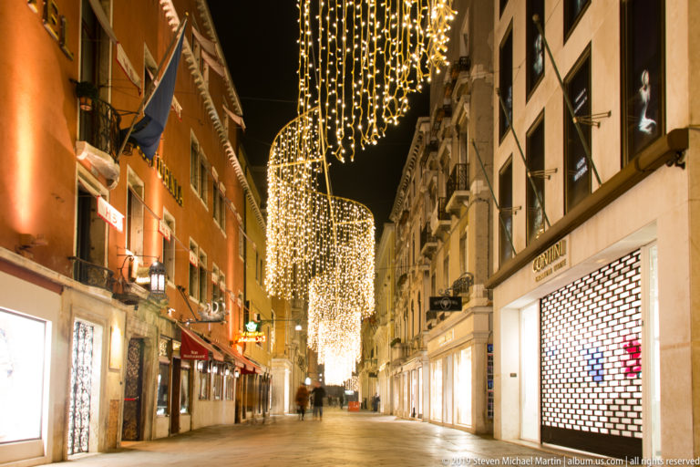 Quiet Venetian shopping street lined with illuminated storefronts beneath sweeping curtains of warm white festive lights.