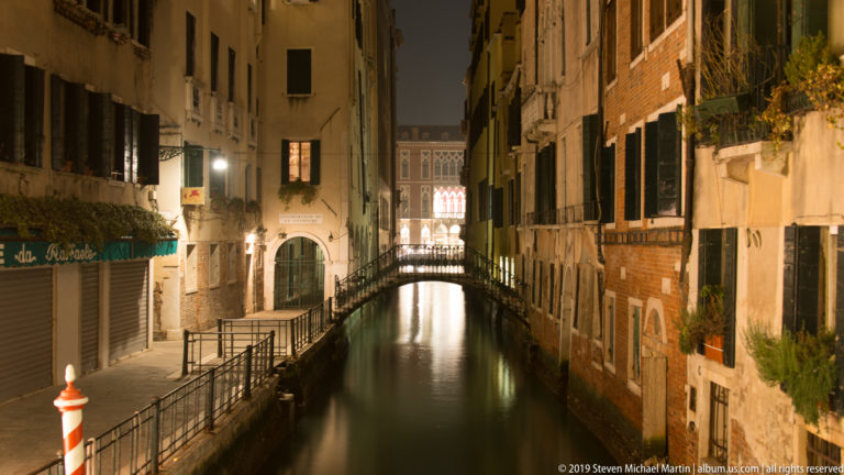Narrow Venetian canal between brick and stucco buildings, with railings, reflections, and a small arched footbridge ahead.
