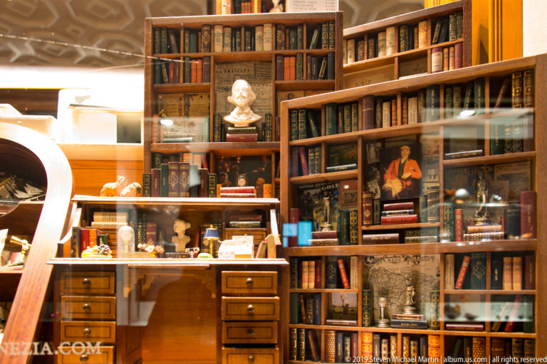 Book-filled shop window with wooden desk, shelves, small sculptures, framed works, and street reflections at night.
