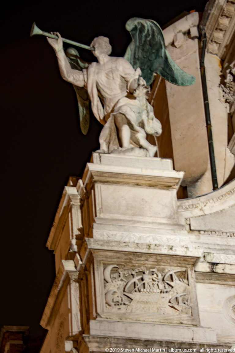Close upward view of pale stone rooftop figures and carved architectural details on Santa Maria del Giglio at night.