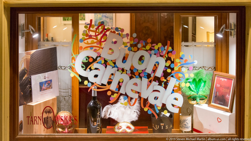 Shop window with large colorful 'Buon Carnevale' lettering, bottles, boxes, masks, and festive decorative merchandise.