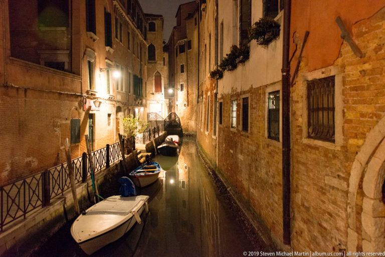 Narrow nighttime canal with several small boats, iron railings, and weathered brick and stucco residential façades.