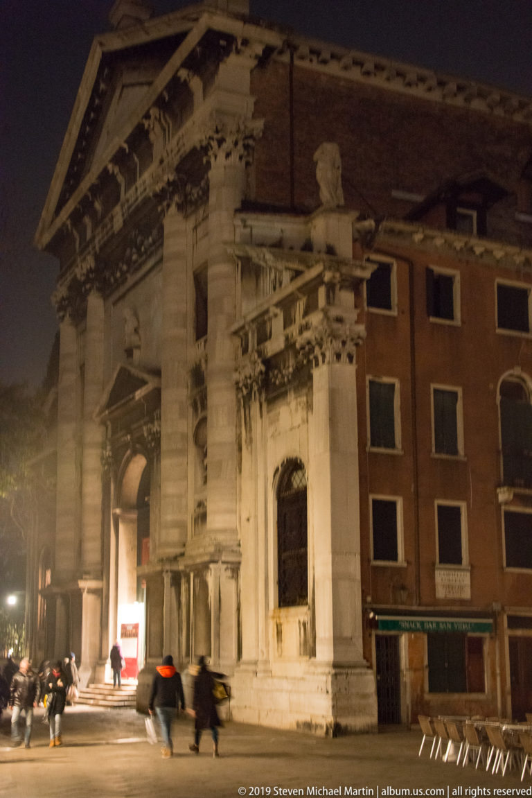 Large illuminated stone church portal with giant columns and arched entrance beside brick buildings and pedestrians at night.
