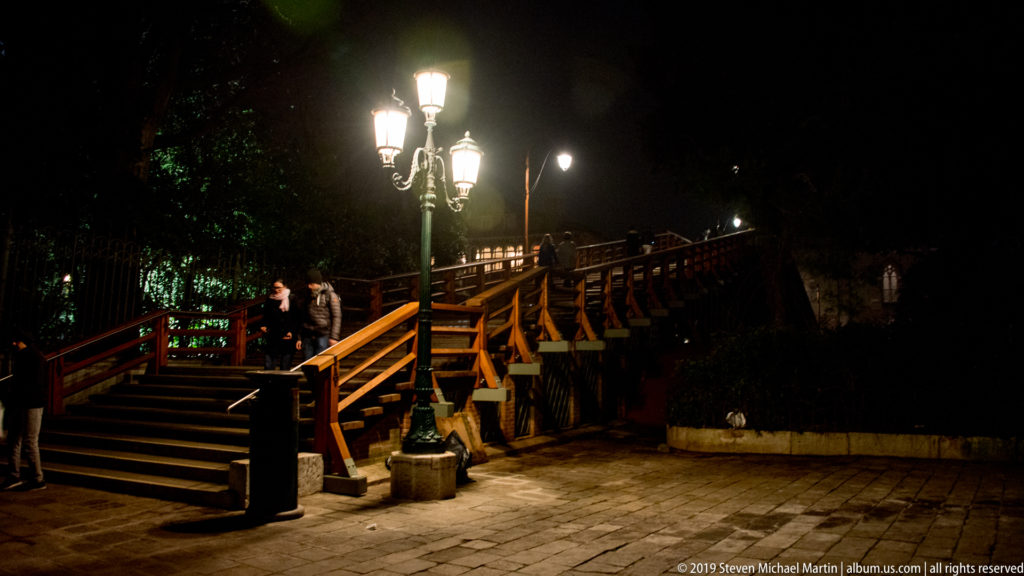 Wooden pedestrian bridge with broad steps, timber railings, ornate streetlamps, and several people crossing at night.