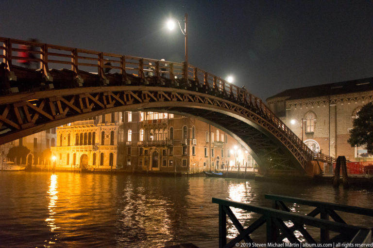 Accademia Bridge arching over the Grand Canal at night, with pedestrians above and illuminated palazzi reflected in the water.