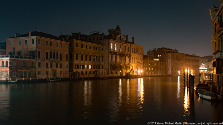 Historic palazzi along the Grand Canal at night, with glowing windows and elongated reflections across dark water.