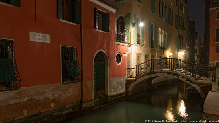 Rio de San Moisè at night beside red and ochre façades, with green shutters and a small arched footbridge.