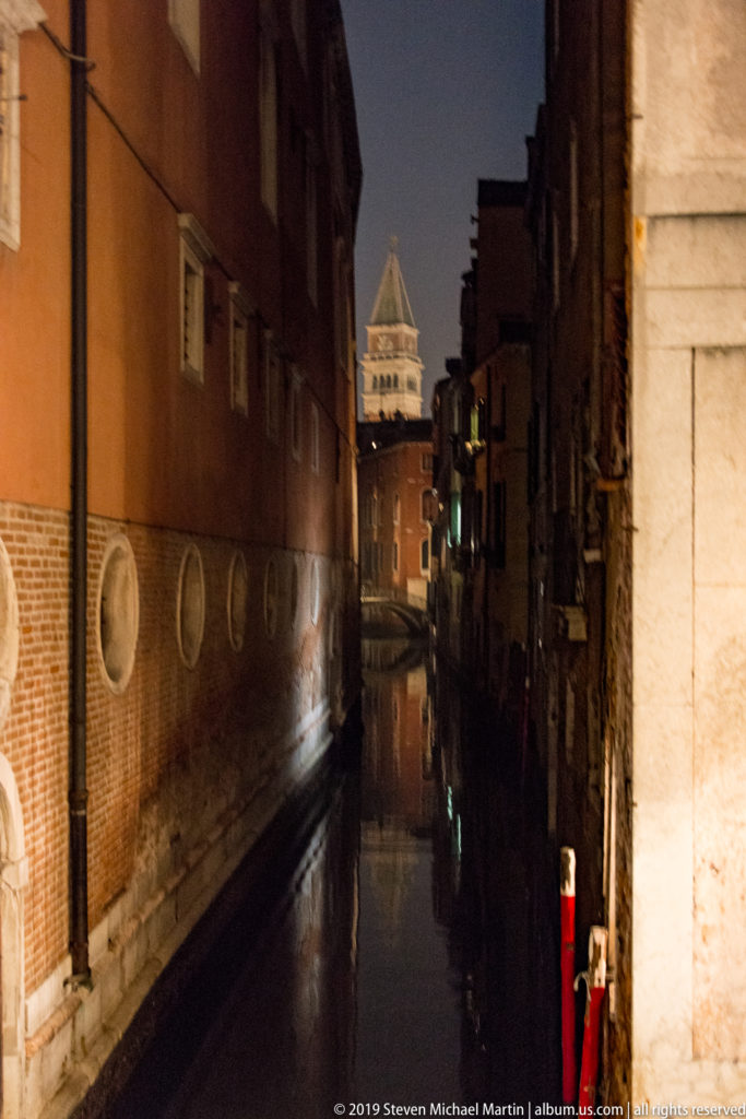 Narrow dark canal between brick buildings leading toward an illuminated bell tower in the distance.
