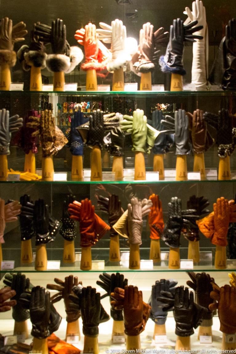 Rows of black, brown, red, white, and colored leather gloves displayed on hand forms in a brightly lit shop window.