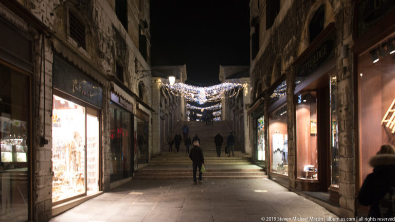 Pedestrians walking along a narrow Venetian shopping passage with bright storefronts and overhead festive lights at night.