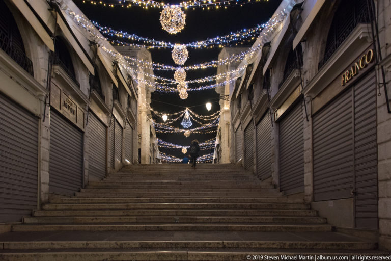 Broad stone steps between shuttered shops beneath repeated swags and medallions of white festive lights at night.