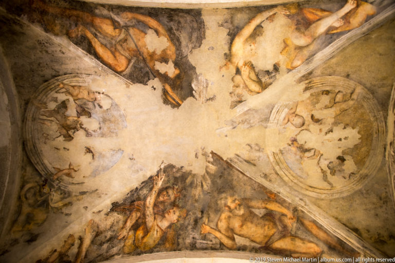 Upward view of an aged frescoed vault with figurative medallions, painted figures, architectural ribs, and areas of worn plaster.