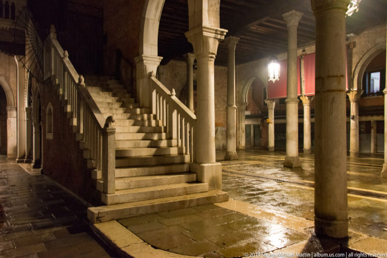 Stone staircase rising through a dim columned interior with arcades, timber ceiling, worn paving, lamps, and red wall hangings.