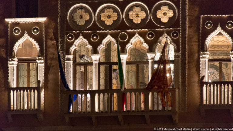 Illuminated Venetian Gothic façade with pointed windows, quatrefoils, balcony balustrade, slender columns, and several hanging flags at night.