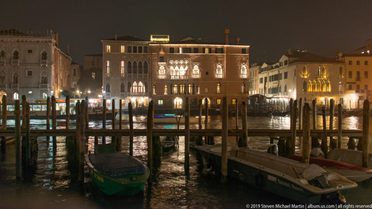 Boats and timber mooring posts at a Grand Canal landing under nighttime illumination in Venice.