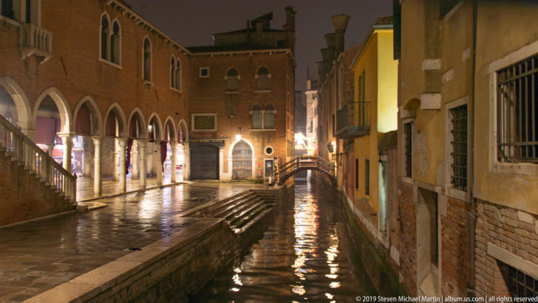 Narrow canal beside an arcaded building and footbridge under nighttime illumination in Venice.