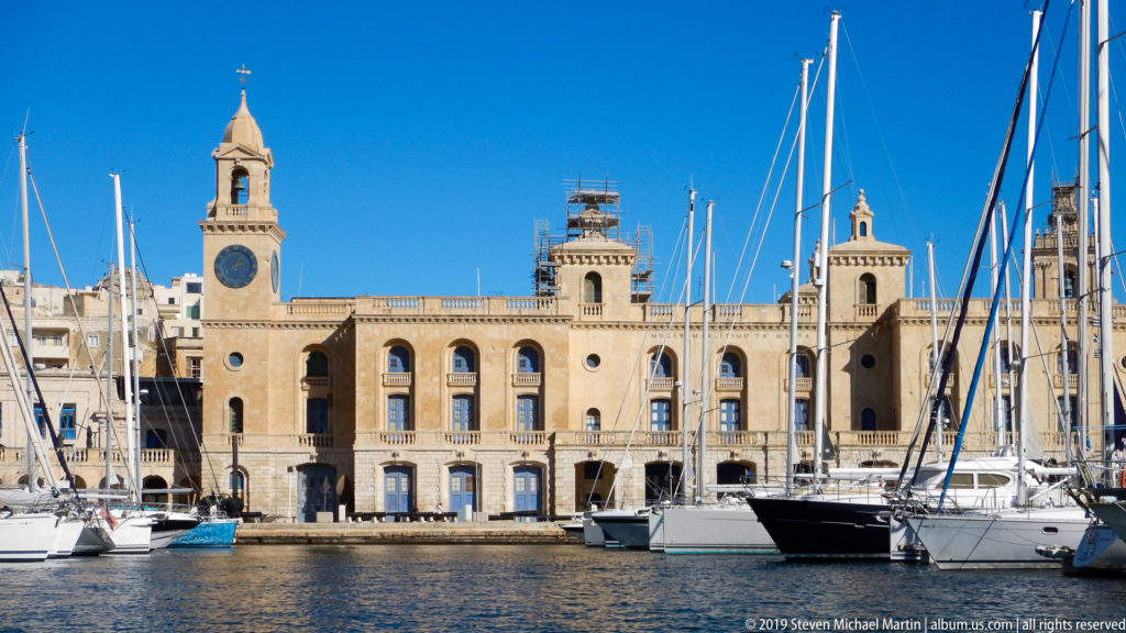 Malta Maritime Museum with central clock tower behind sailboats and tall yacht masts in Vittoriosa marina.