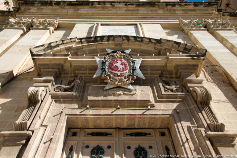 Low-angle view of an ornate limestone doorway crowned by a red-and-white heraldic crest on an eight-pointed cross.
