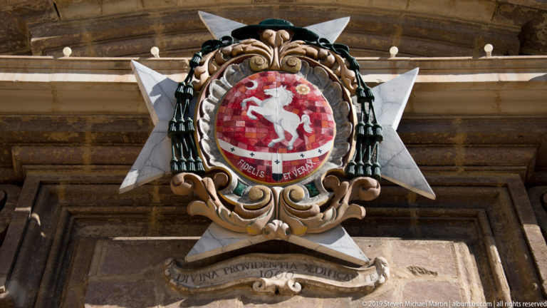 Ornate crest with red shield, white rearing horse, carved scrollwork, and a large white eight-pointed cross behind it.
