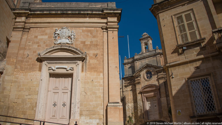 Two limestone buildings with monumental doorways, heraldic carving, classical surrounds, and a circular window beneath a blue sky.