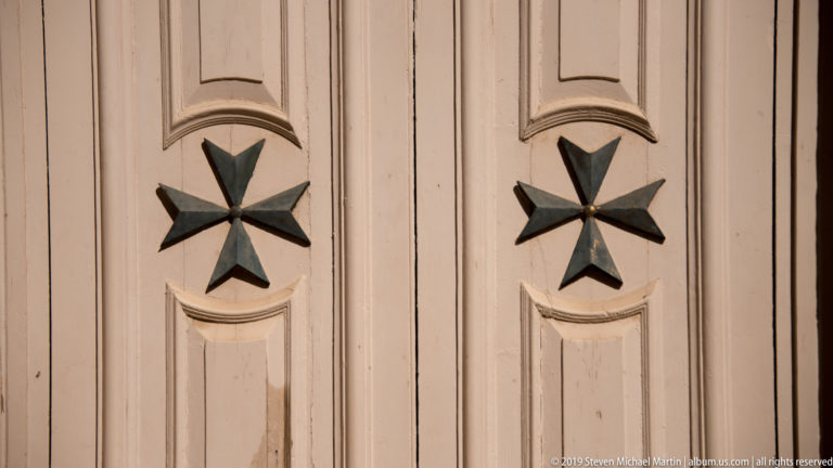 Two dark eight-pointed Maltese crosses mounted on pale paneled wooden doors with curved molding.