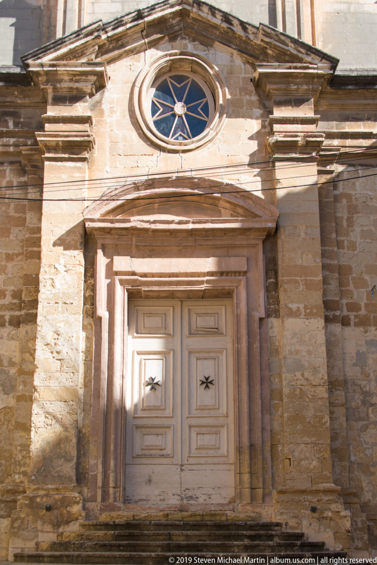 Paneled double door with Maltese-cross motifs beneath a circular radial window and triangular pediment in a limestone façade.
